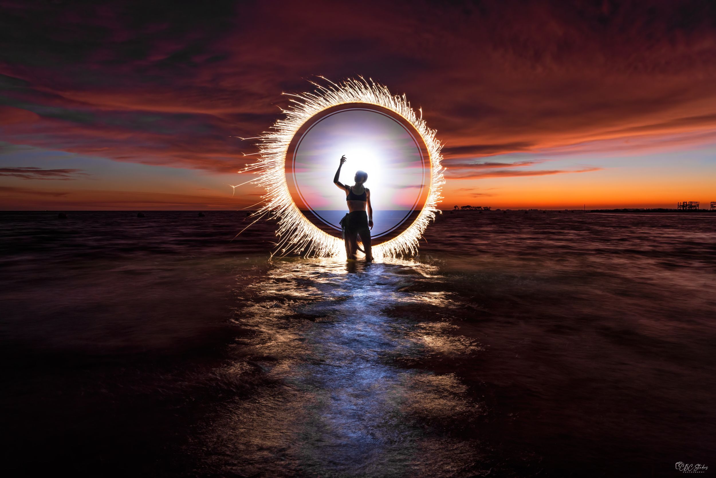 Silhouetted figure stands against dramatic sunset sky creating circular solar halo effect over reflective water surface.