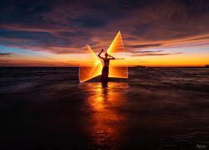 A silhouette against a sunset sky creates light paintings shaped like triangles while standing in shallow coastal waters.