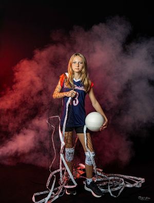 Volleyball player in navy uniform posed with net and ball in red smoke effect studio portrait.