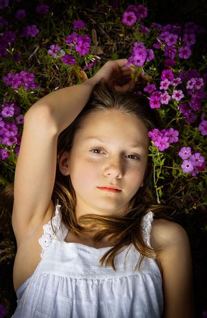 A person in a white dress lies among vibrant purple flowers in a garden setting.