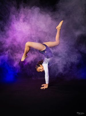 Gymnast performing handstand in purple leotard with dramatic smoke effects in studio setting.