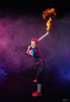 Young performer in colorful outfit holds flaming torch against purple smoky background.