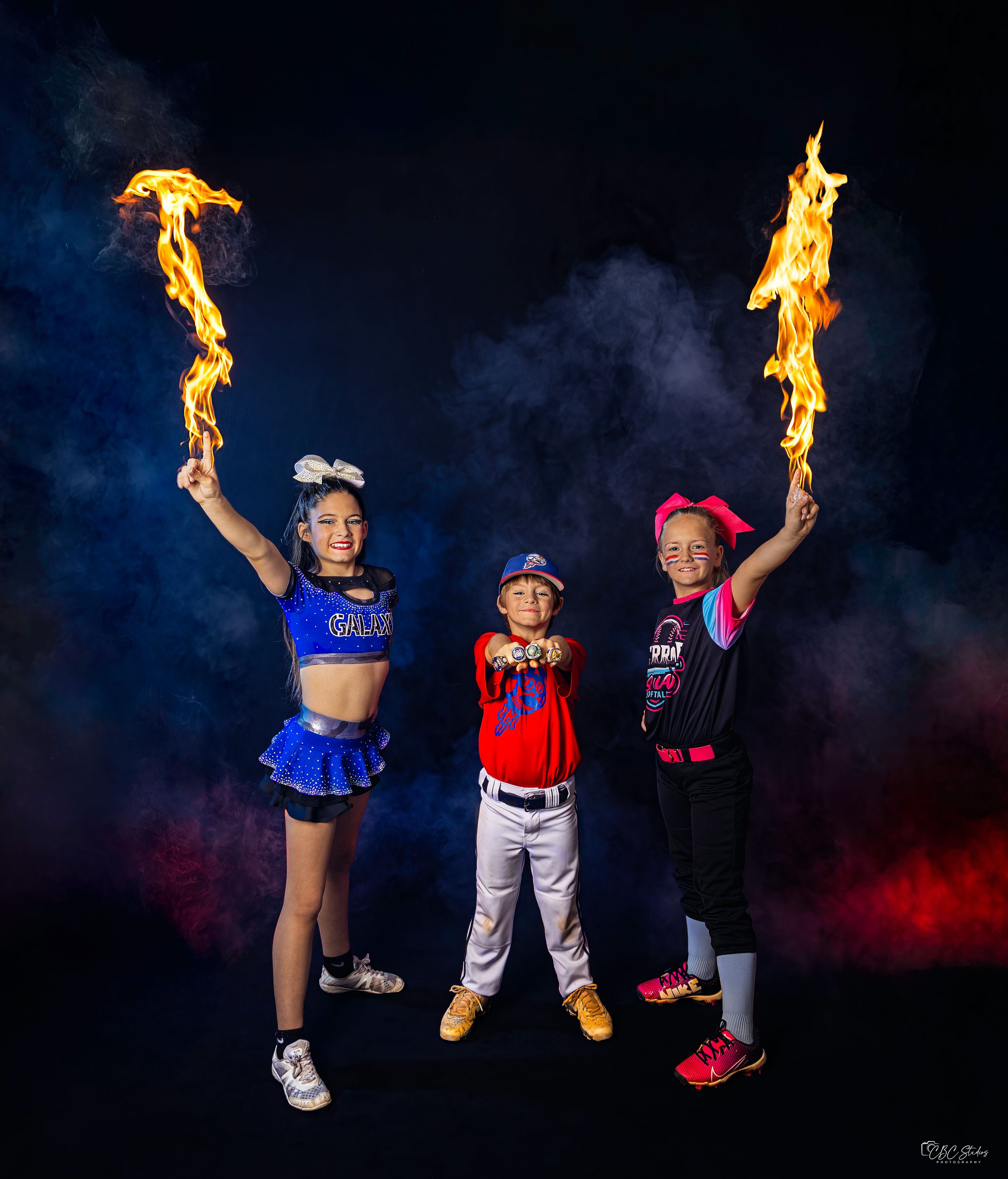 Three performers holding flaming torches pose together against dramatic dark background.