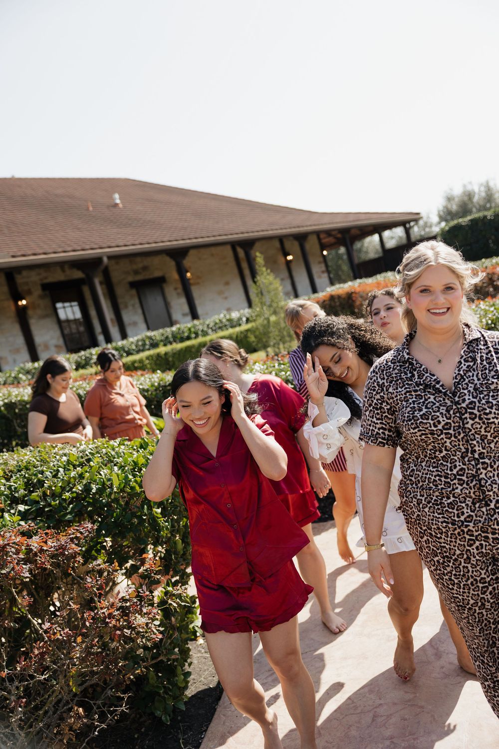 A group of people dancing and laughing together outdoors on a sunny day near a building with hedges and bushes.