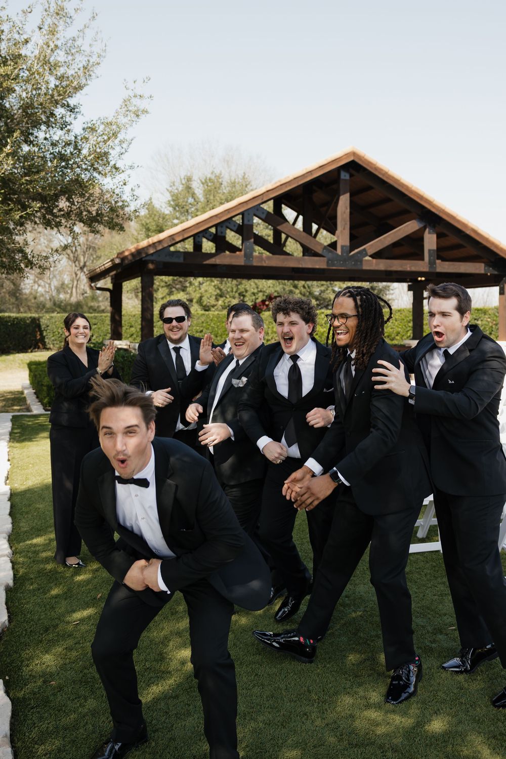 A group in formal black suits and bow ties pose playfully on grass in front of a wooden pavilion structure.