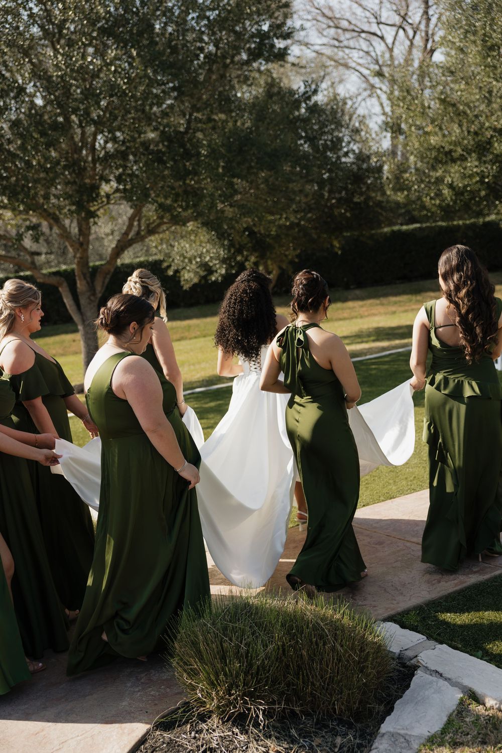 A row of bridesmaids in olive green dresses sit together on stone steps in an outdoor garden setting.