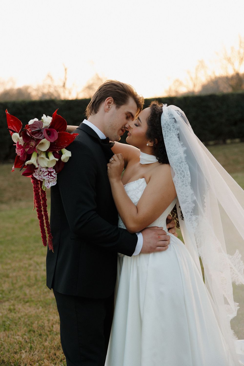 Wedding couple shares an intimate kiss in a field during golden hour with burgundy floral bouquet.