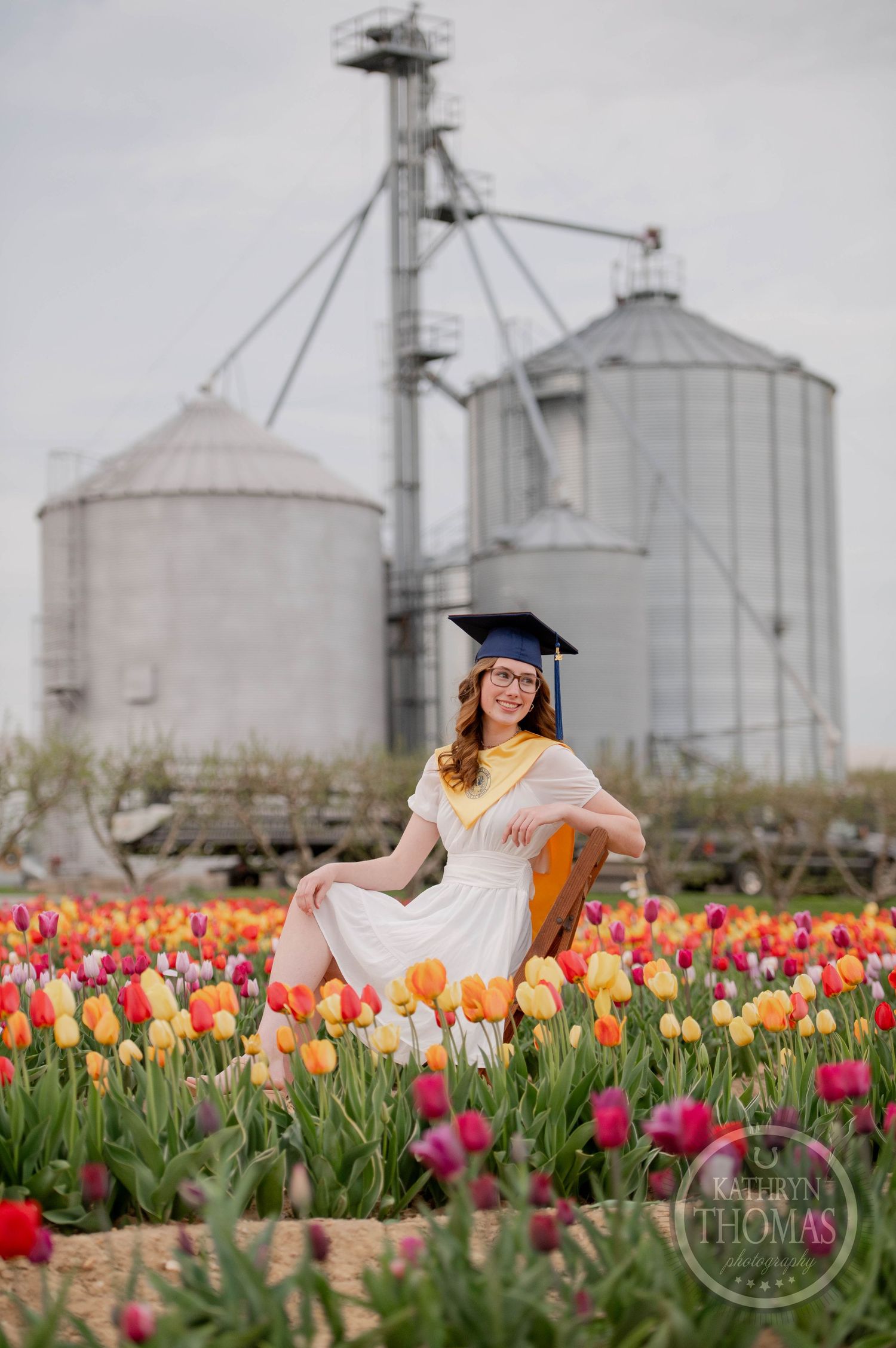 Graduate in white dress poses among vibrant tulip field with farm grain silos in the background.