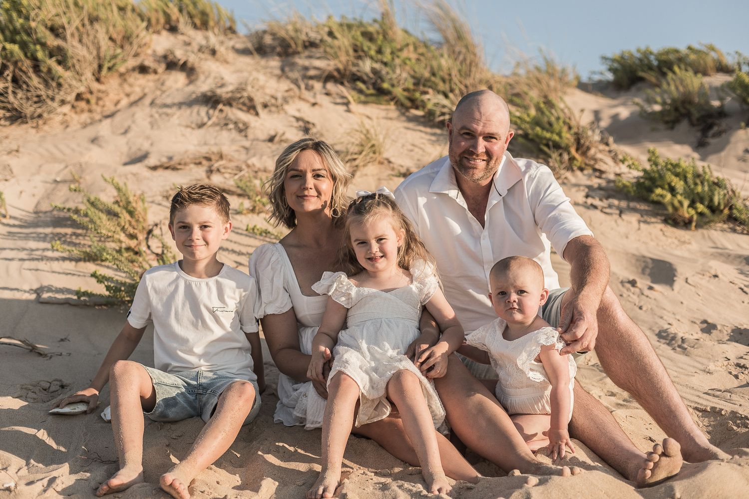 A family sits together on a sandy beach during golden hour wearing coordinated white and neutral colored clothing.