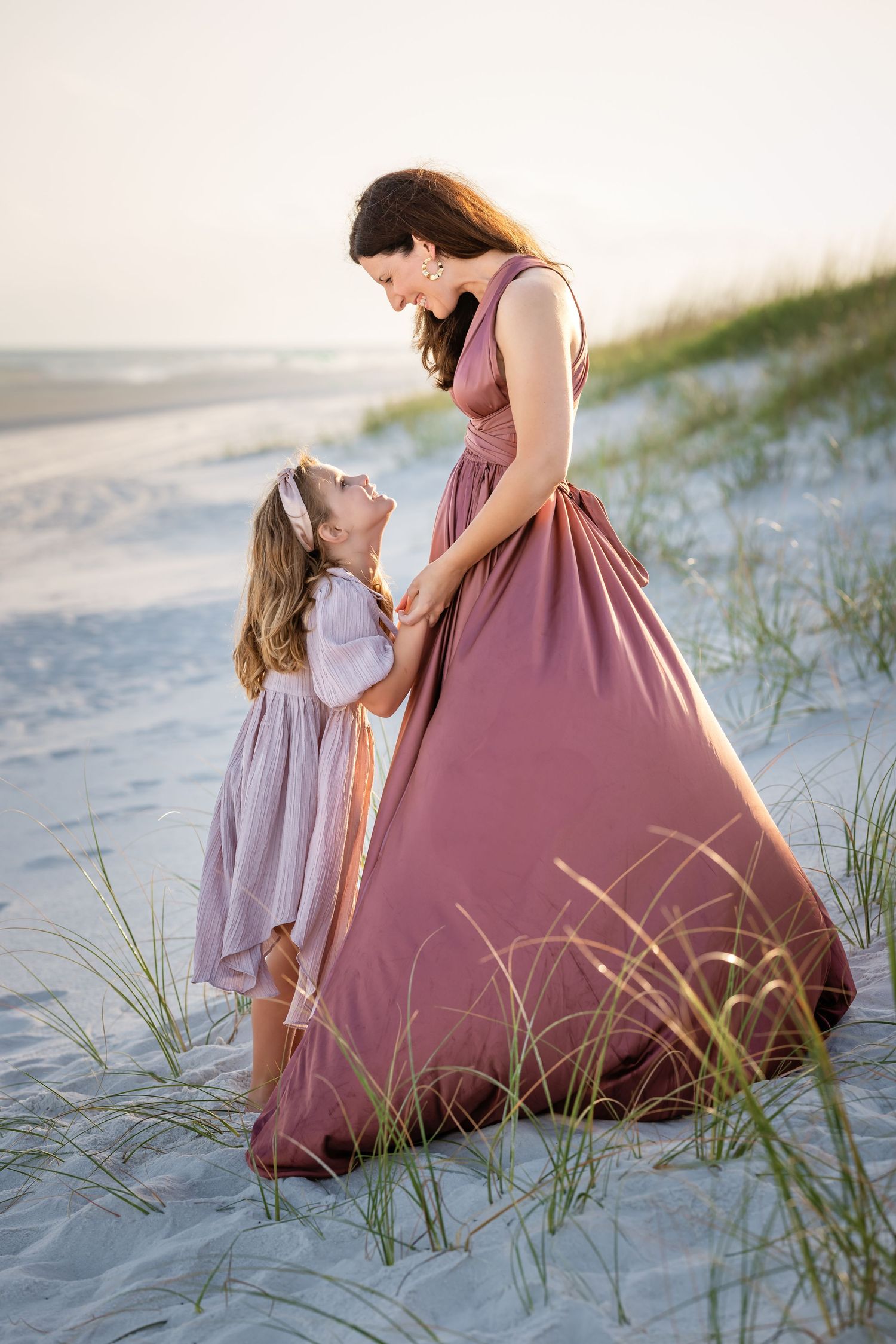 Two people in mauve dresses share a tender moment on a sandy beach with sea grass at sunset.