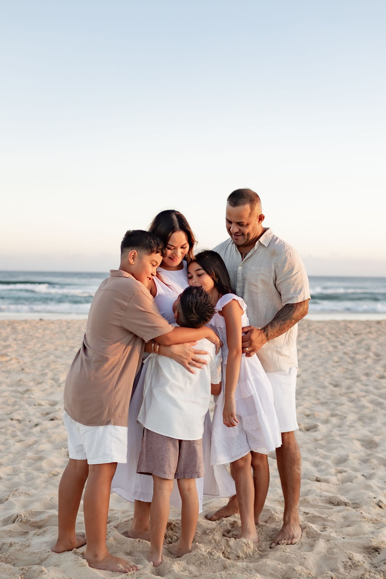 Family wearing white and beige clothing poses together on beach at sunset while young child dances.