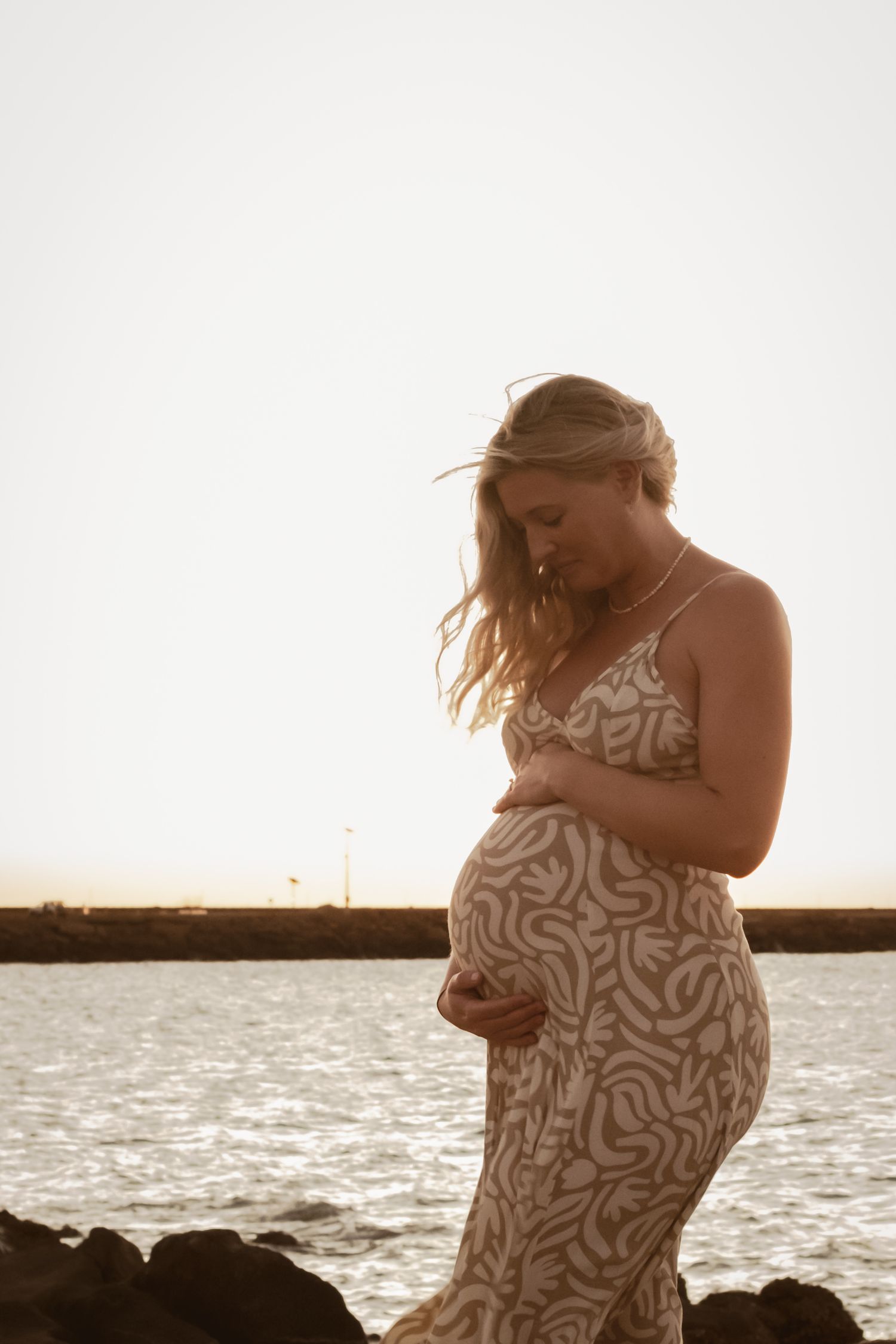 Silhouette of a pregnant woman in a patterned dress standing by the ocean at sunset.
