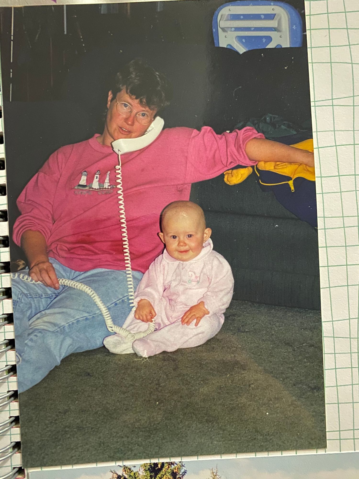 A person in a pink sweatshirt sits against a dark wall with a baby in a light pink outfit.