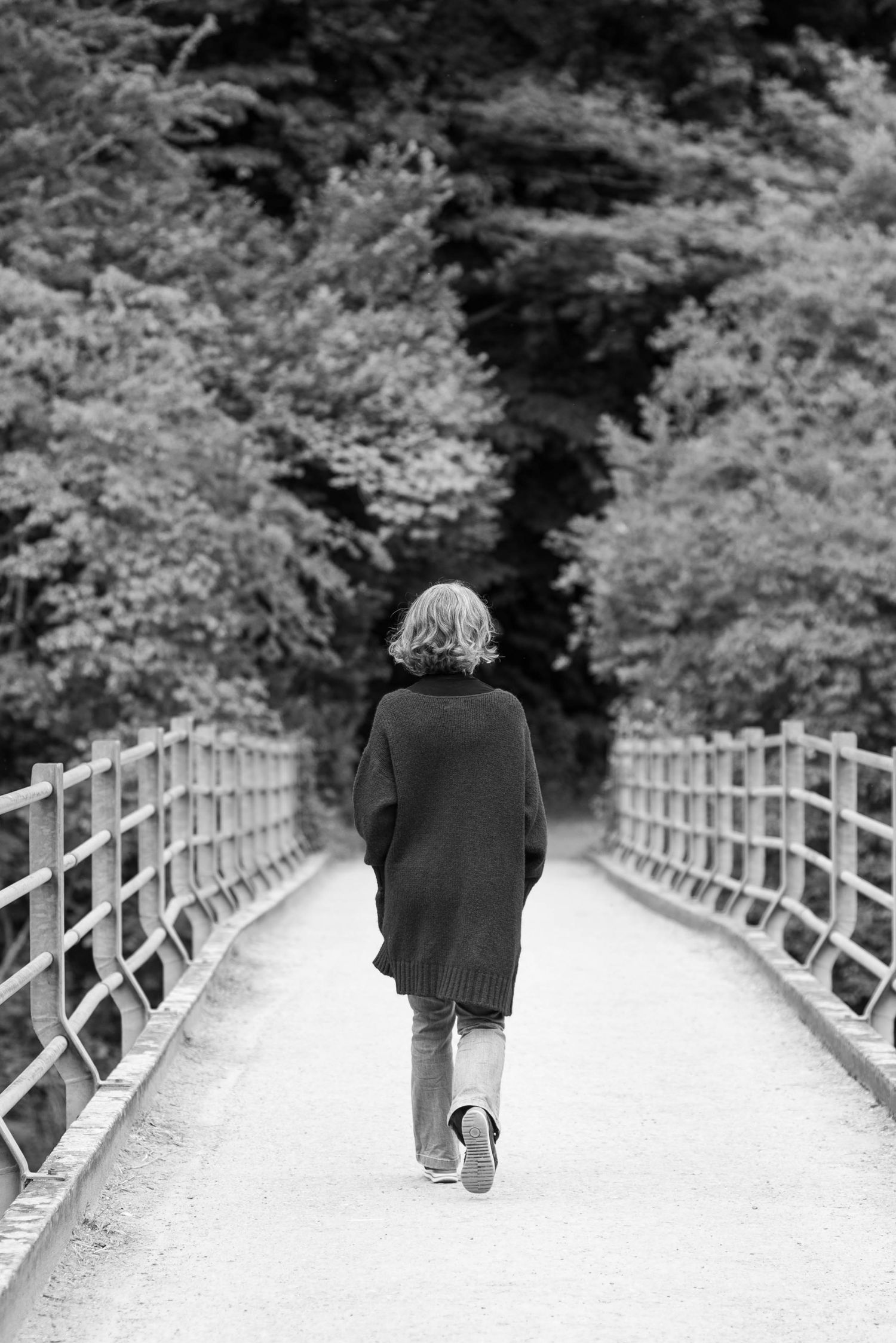 Black and white sequence of someone walking away on a white fenced path surrounded by trees.