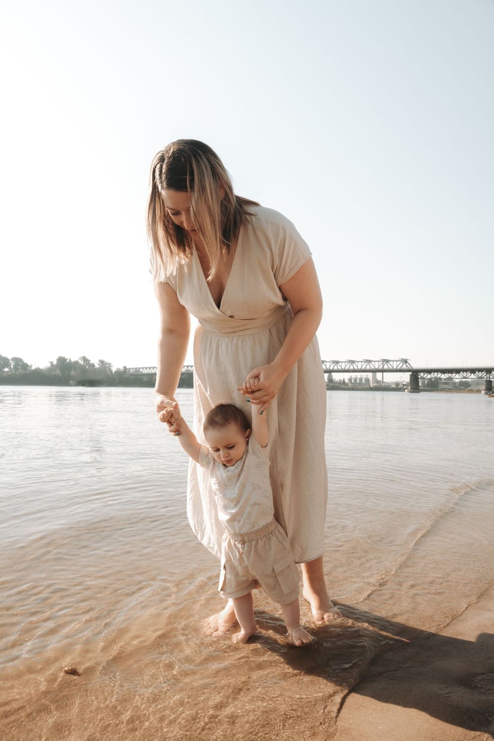 Mother in beige dress helps toddler walk along sandy riverbank shore on a sunny day.