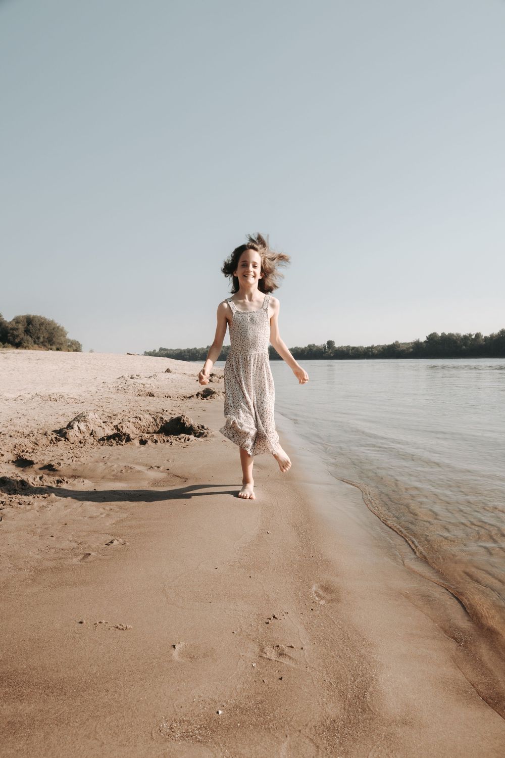A young girl in a white sundress runs barefoot along a sandy beach shoreline on a sunny day.
