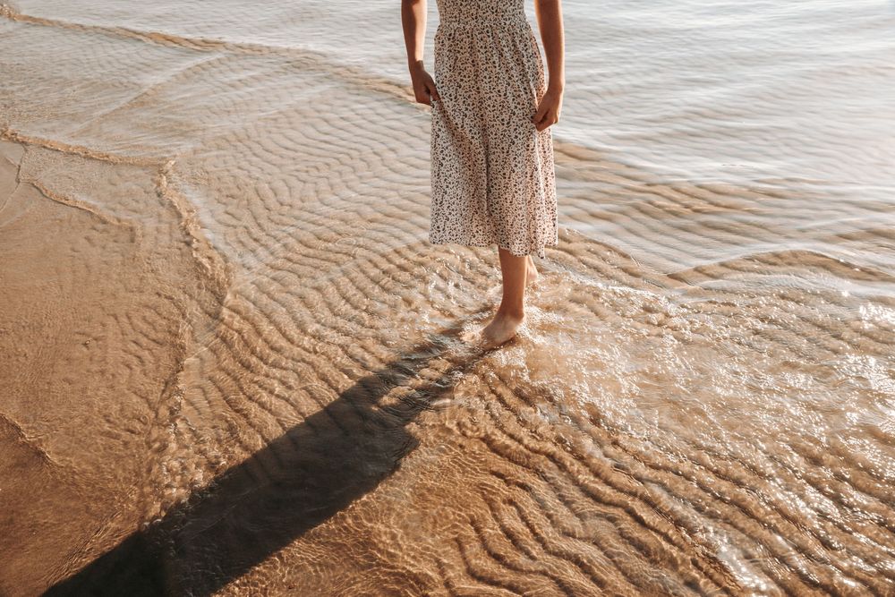 Woman in floral dress standing barefoot on sandy beach shore with gentle waves washing over her feet in golden light.