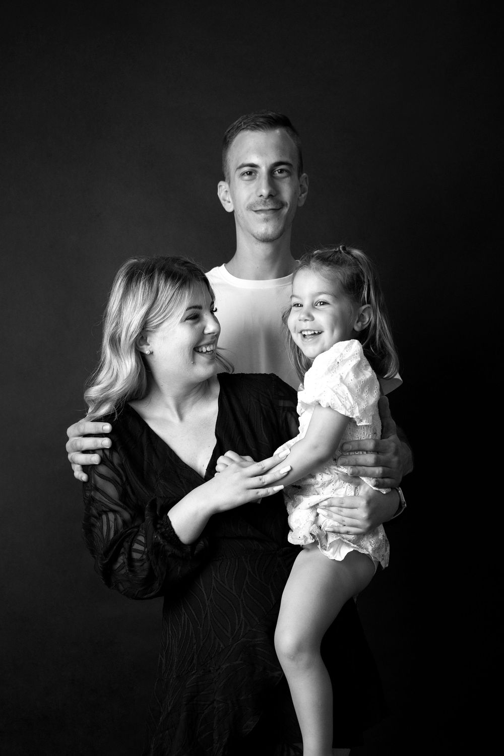 A studio portrait of a family of three in black and white against a dark background.