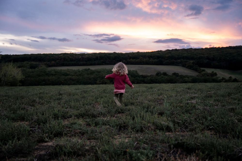 Little girl runs through a field at sunset with dramatic pink clouds in the sky.