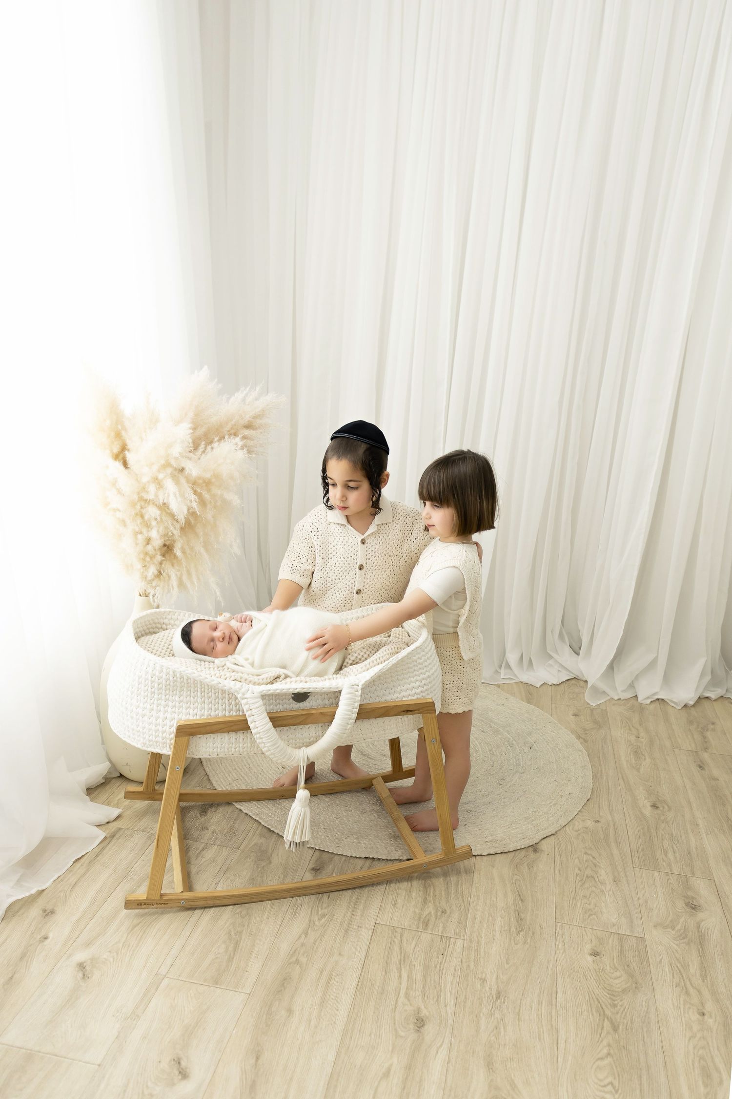 Two siblings lovingly watch over a baby in a white rattan bassinet against a light backdrop.
