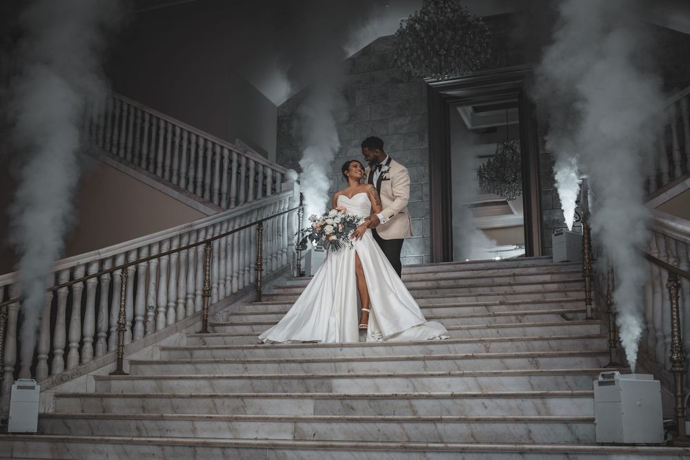 A couple in elegant wedding attire stand on grand stone steps surrounded by dramatic smoke effects at dusk.