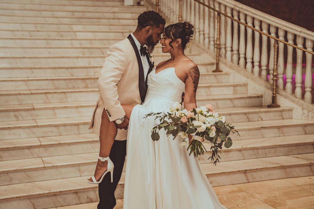 A newly married couple embraces on elegant stone steps as the bride holds a beautiful white floral bouquet.