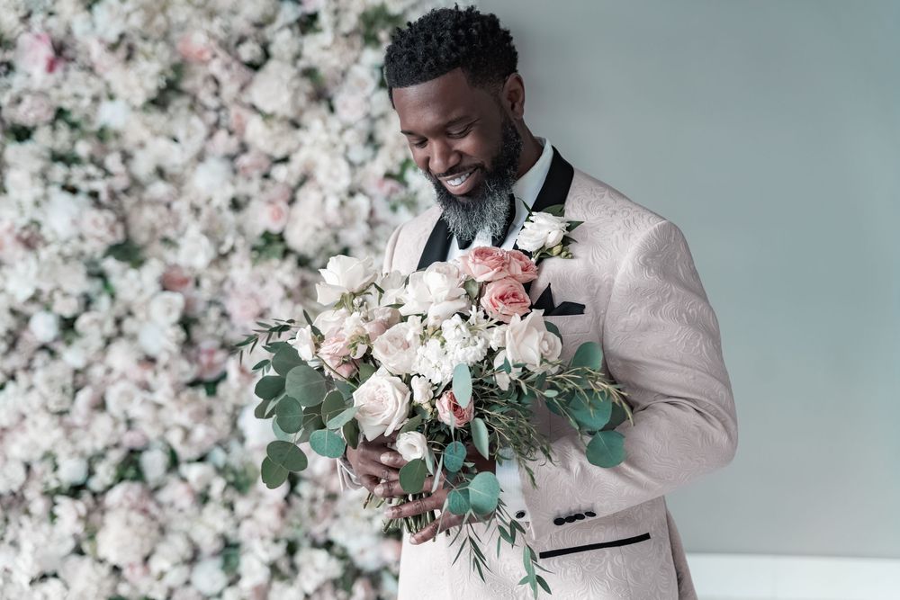Groom in light colored tuxedo smiles while holding a romantic pink and white floral bouquet against flower wall backdrop.