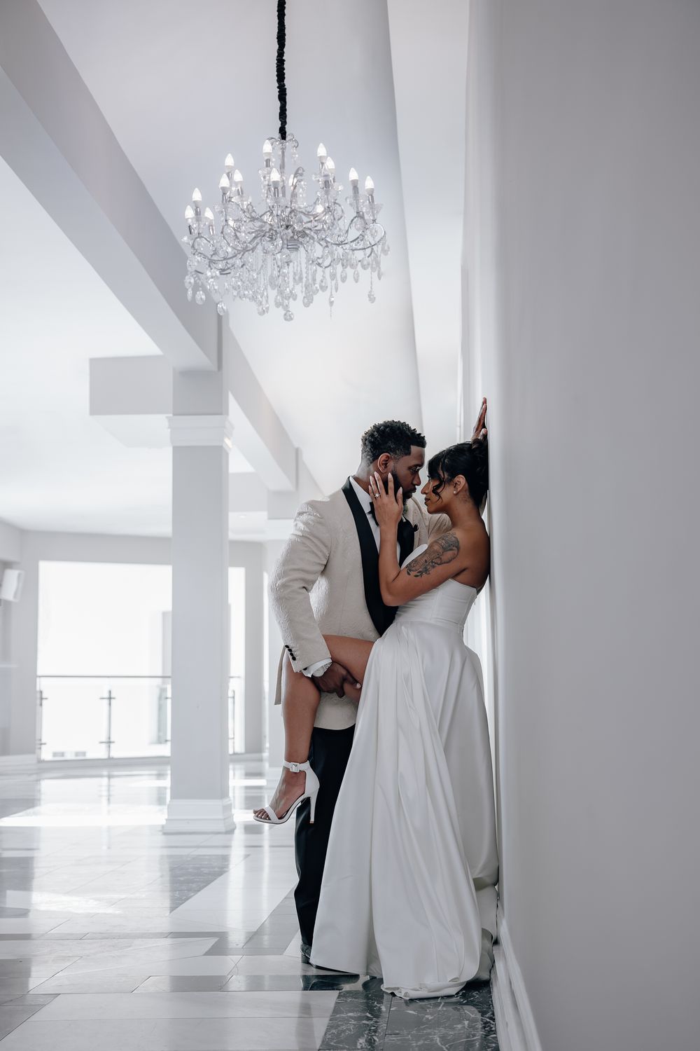 Elegant wedding couple embraces in a white hallway under a crystal chandelier in a bright modern venue.
