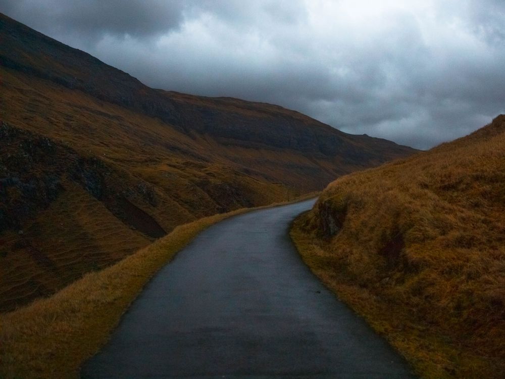 A winding wet road curves through moody autumn hills under dark storm clouds in the Faroe Islands
