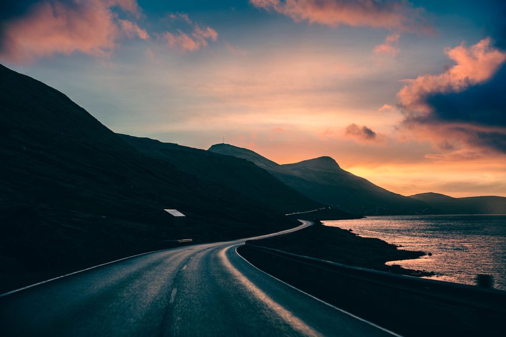 Winding coastal road curves along dark mountains at sunset with dramatic pink and orange clouds over the ocean.