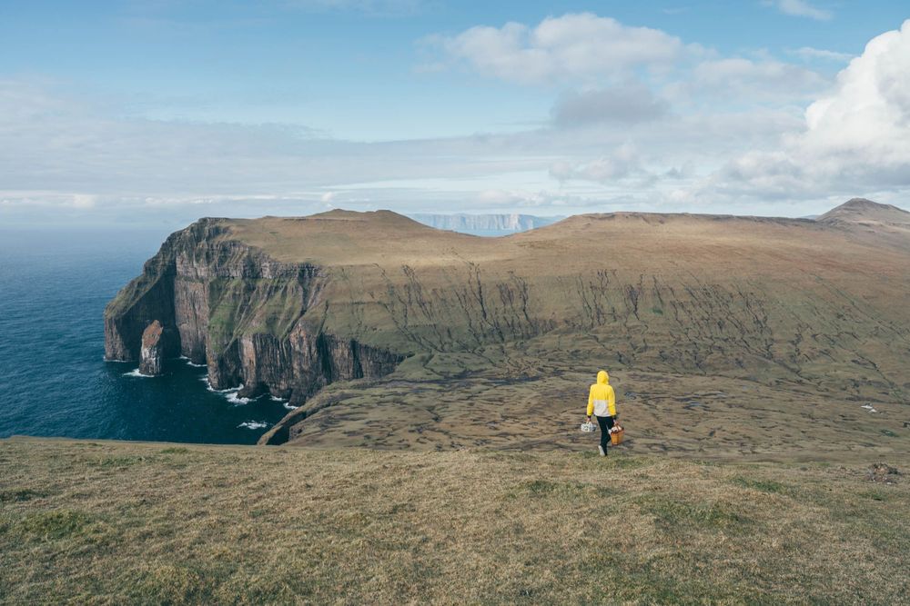 Person in yellow jacket stands on grassy cliff overlooking dramatic coastal cliffs and ocean in the Faroe Islands.