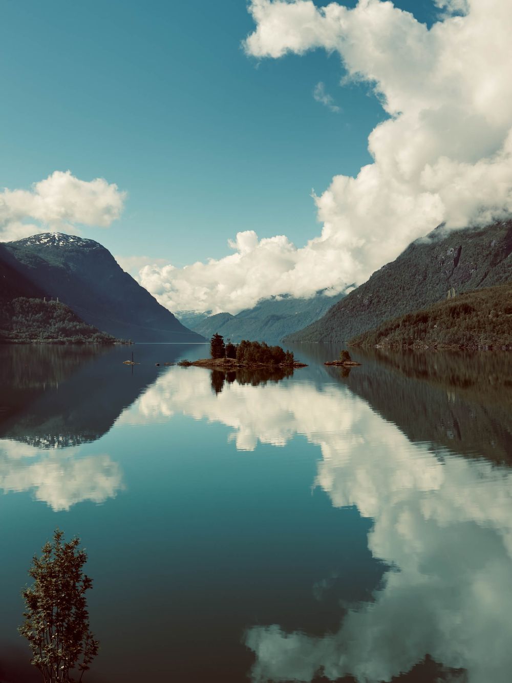 Tranquil mountain lake with small forested island reflecting in mirror-like water beneath cloudy blue skies in Norway.