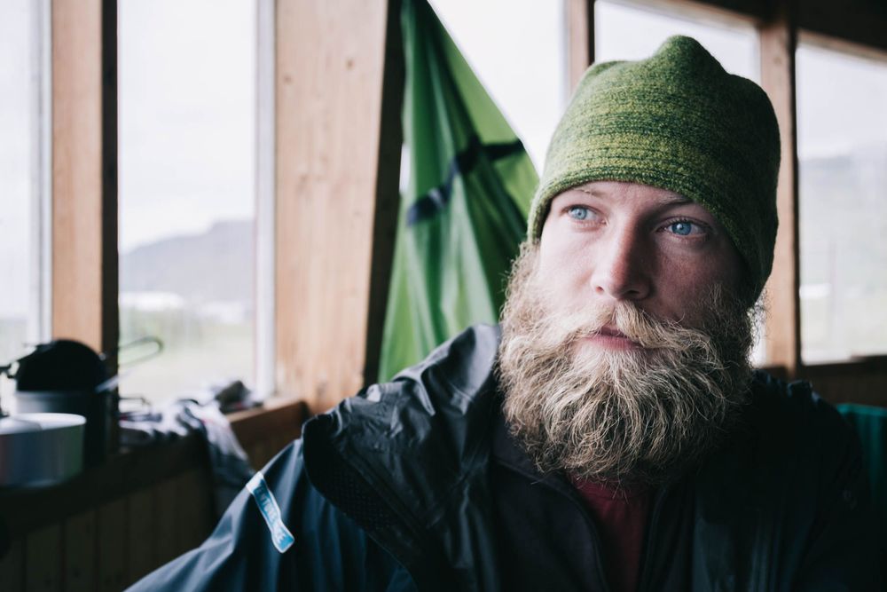Portrait of some nordic person wearing a green winter hat with a full beard sitting near a window.