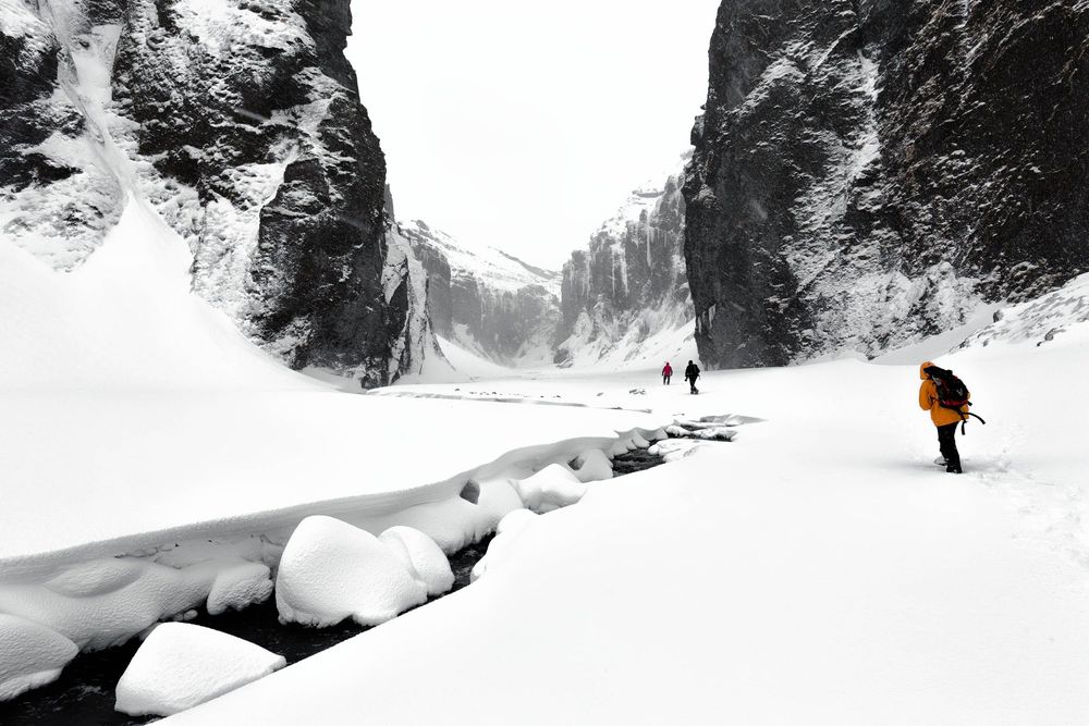 Winter adventurer in orange jacket hiking through a dramatic Icelandic snow-filled mountain canyon with towering cliffs.