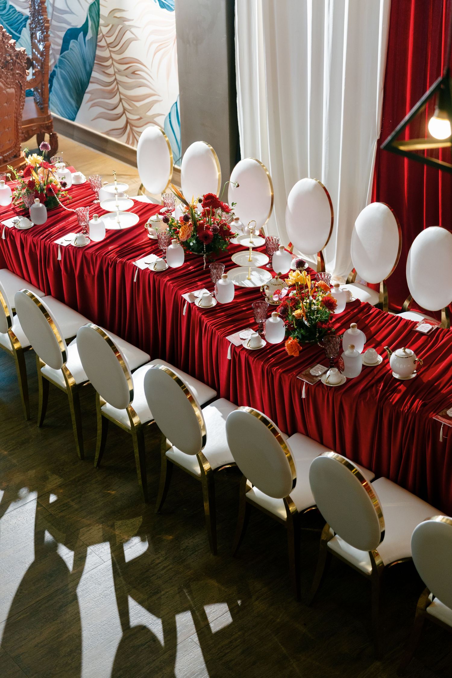 An elegant wedding reception table with red tablecloth, white chairs, and colorful floral centerpieces arranged in a long line.