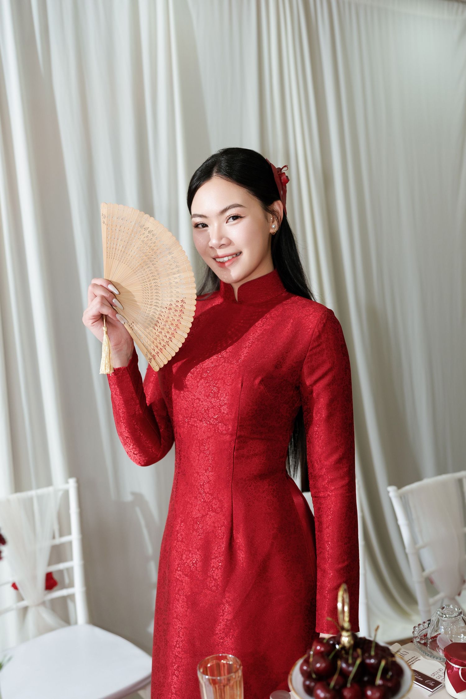 Someone wearing a traditional red Ao Dai dress holds a wooden fan while standing in an elegant party setting.