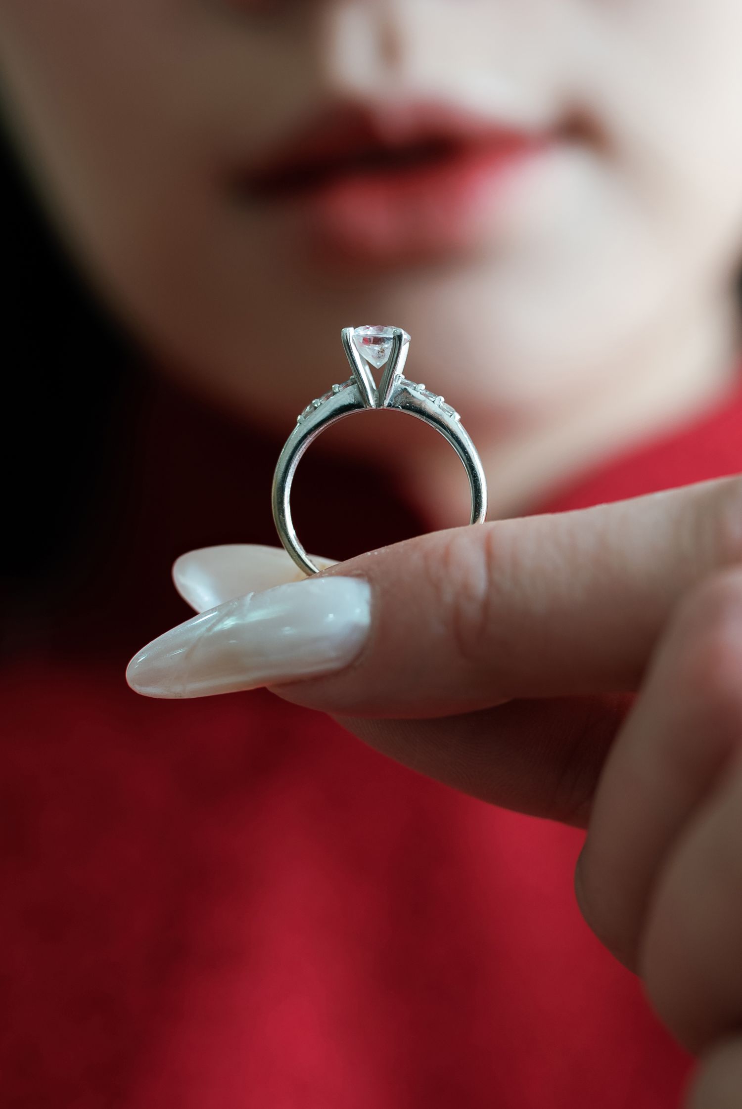 A hand wearing red holds up a silver diamond engagement ring against a blurred background.