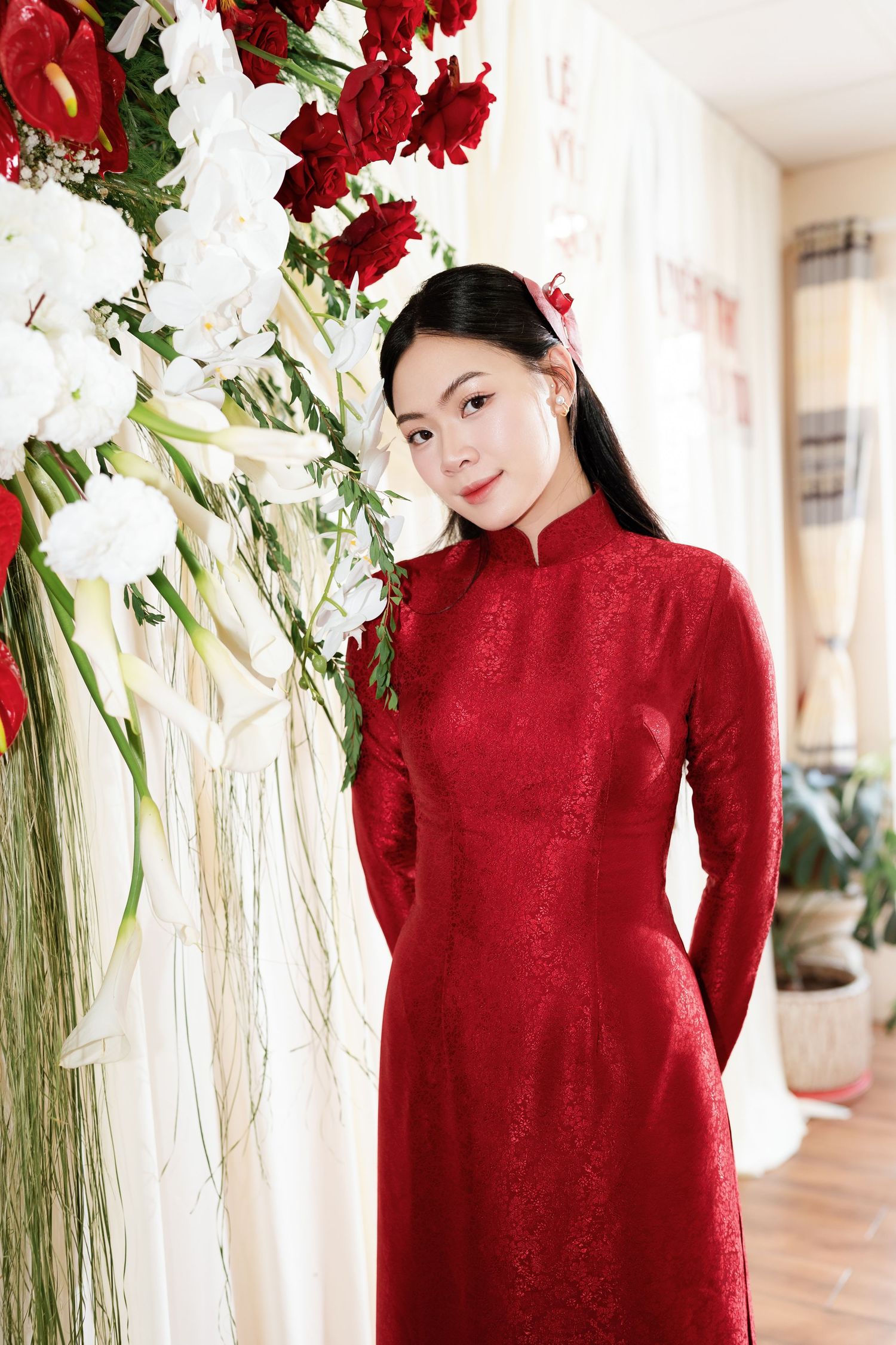 Traditional red ao dai dress with ornate details stands beside elegant floral arrangement of red and white blooms.