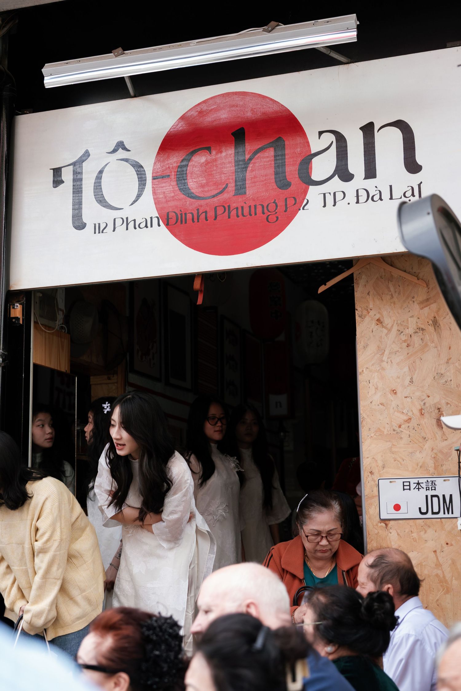 People gather outside a Japanese restaurant with red sun logo and 'To-Chan' signage on a busy street corner.
