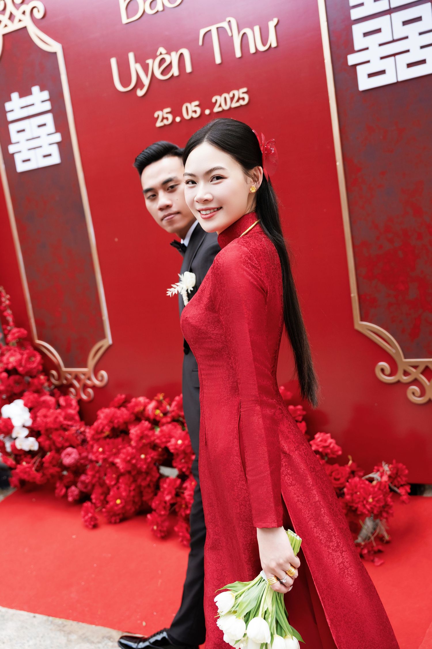 Couple in traditional wedding attire posing against a red backdrop with floral decorations and Chinese characters.