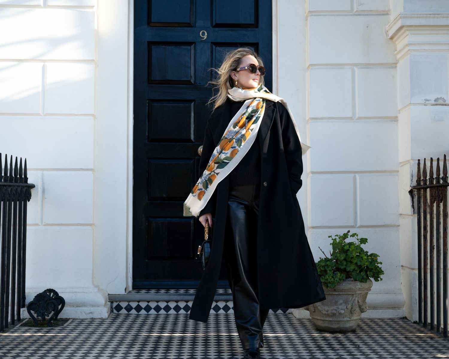 Stylish woman in black coat and colorful scarf poses outside elegant London townhouse.