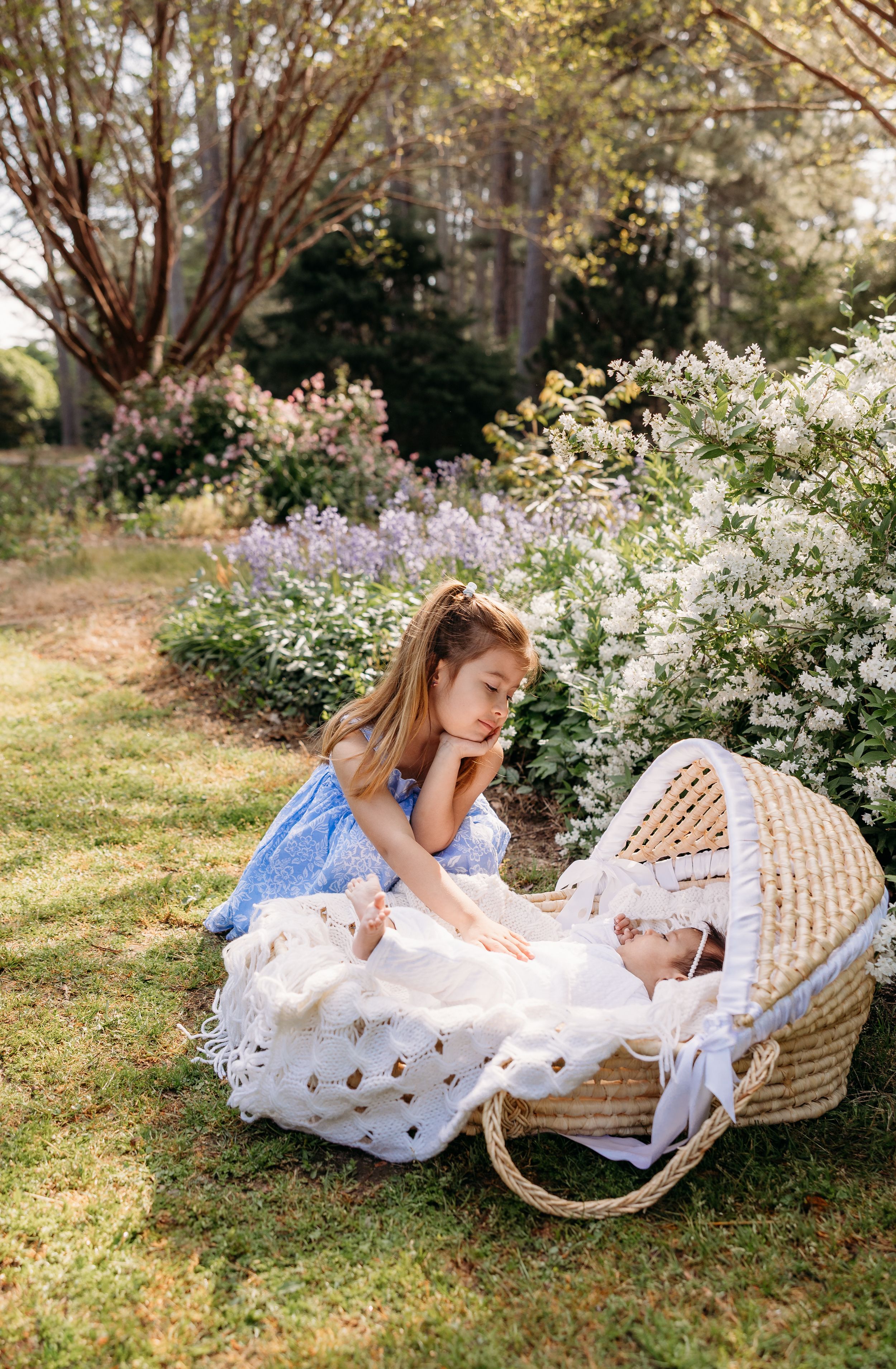A young girl in a blue dress gazes lovingly at a newborn baby in a wicker Moses basket outdoors.
simple timeless natural