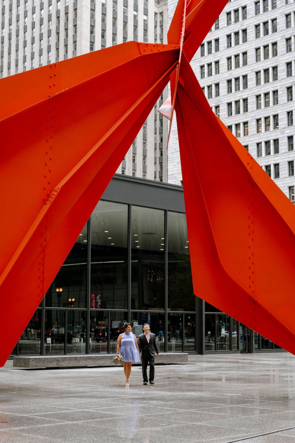 A couple walks beneath a towering red abstract sculpture against a modern glass building and skyscraper backdrop.