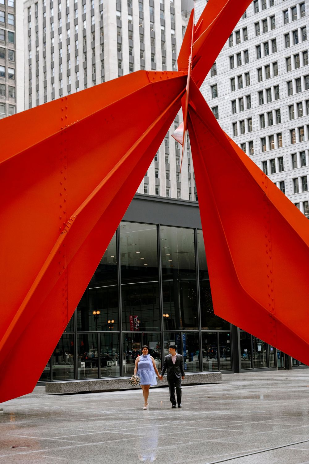 Two people walk beneath a striking red architectural sculpture outside a modern glass building in Chicago.