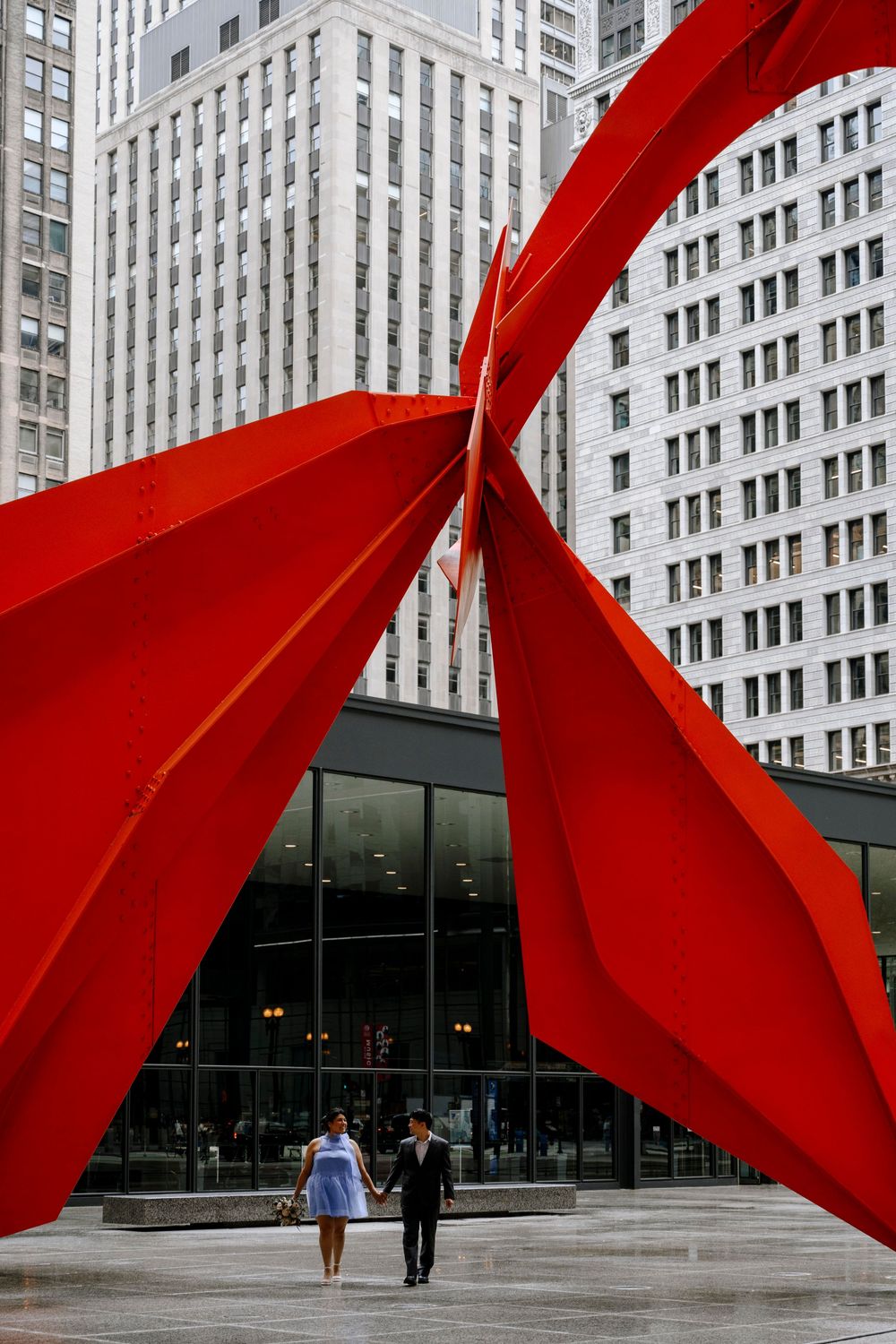 A couple walking under a large red metal sculpture in front of a modern urban building.
