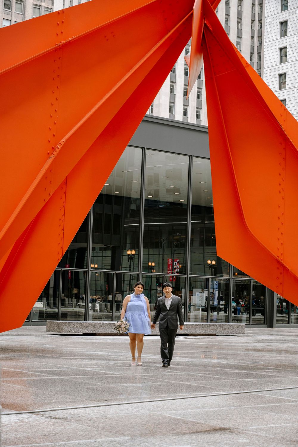 Couple strolling under large red geometric sculpture in front of modern glass building.
