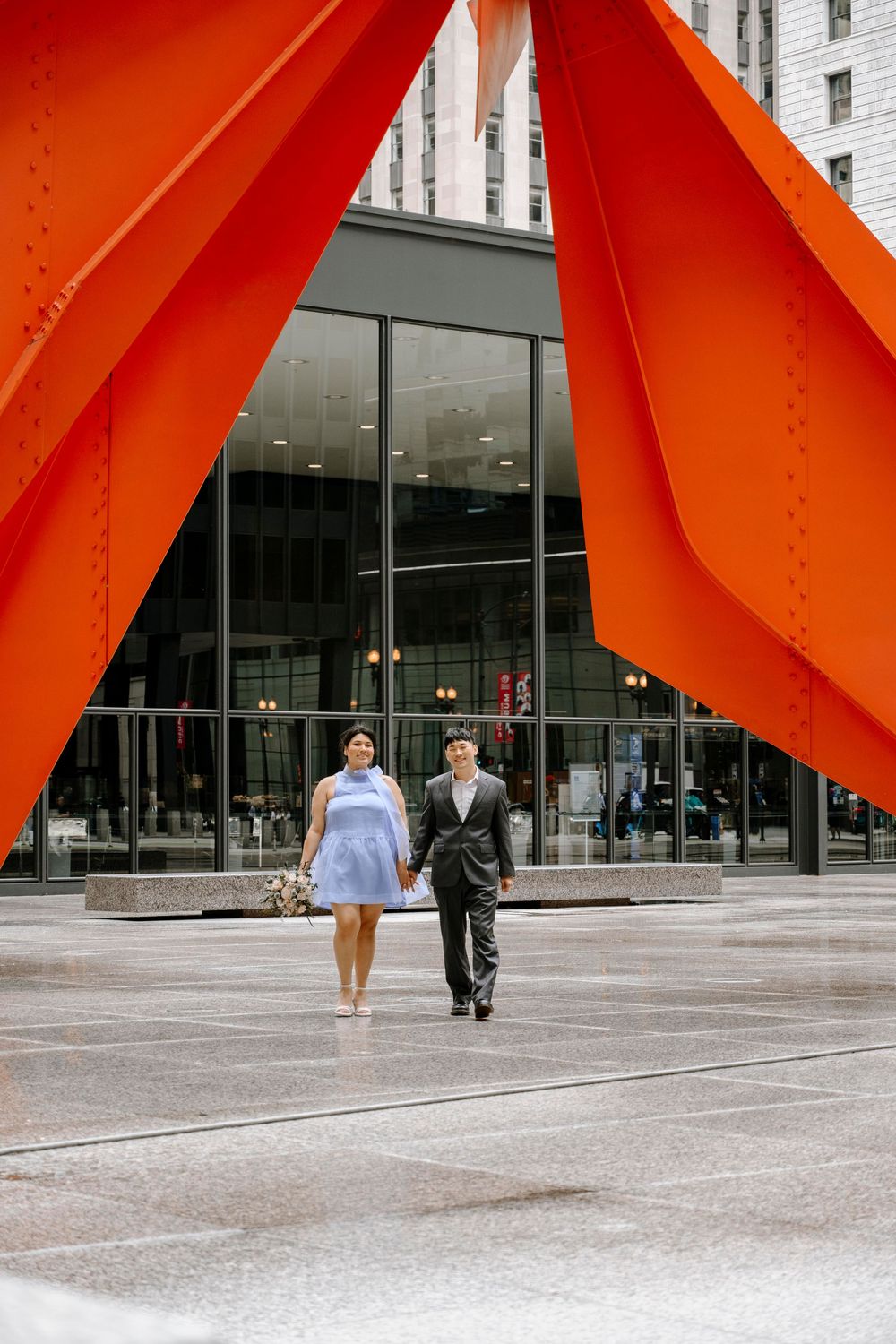 Two people walk beneath a large orange sculpture in front of a modern glass building in Chicago.