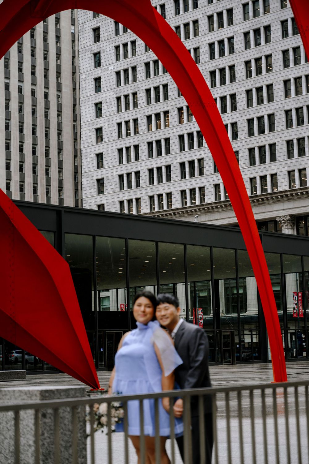 A couple poses beneath a towering red sculpture arch in front of a modern urban building.