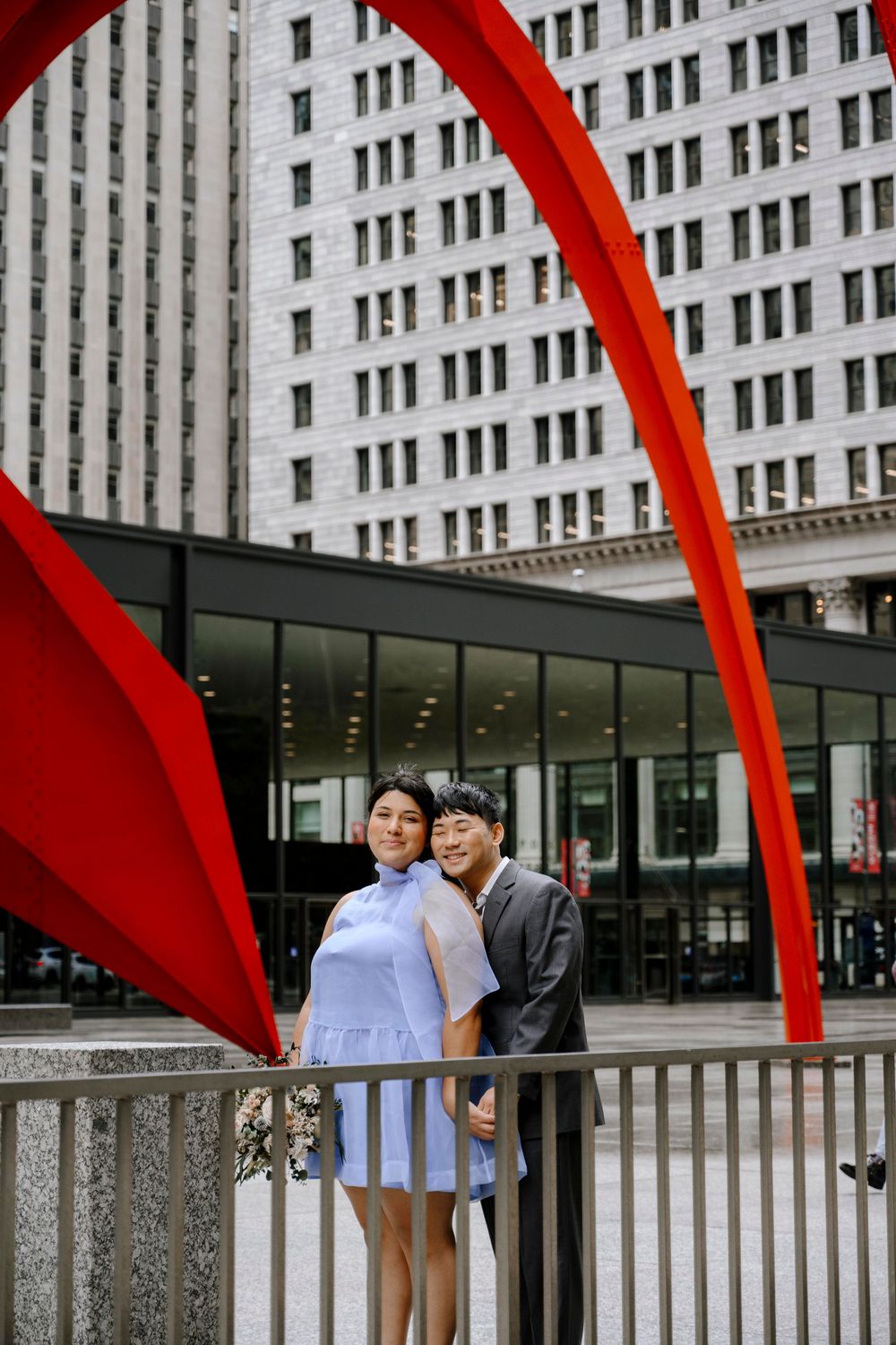 A couple poses under a vibrant red sculpture arch in front of a modern urban building.