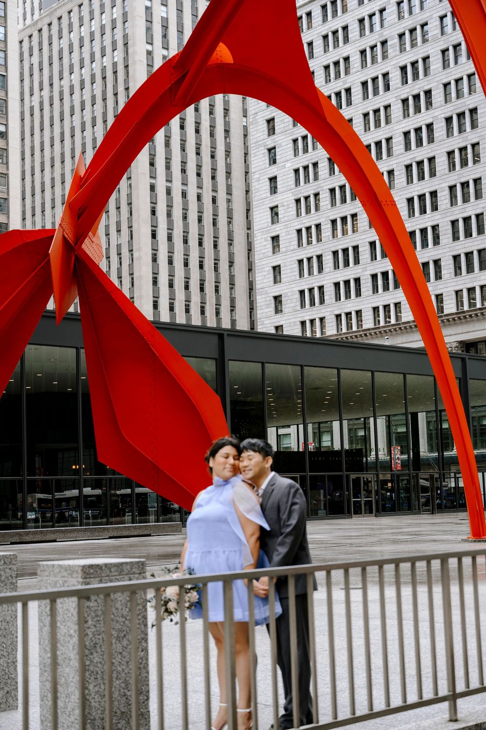 A couple shares an intimate moment under a large red metal sculpture in front of an urban building.